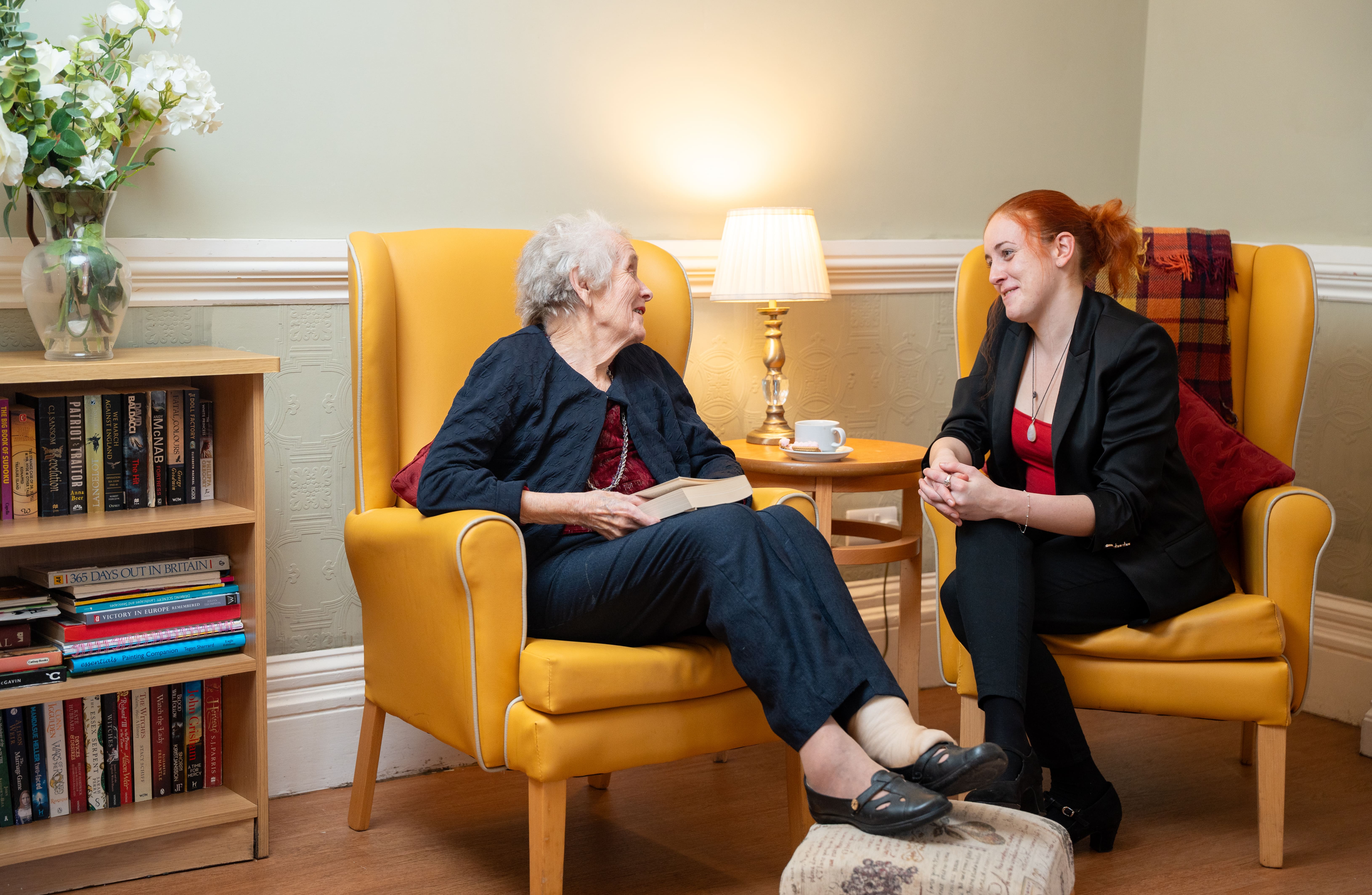 An elderly resident chats with a younger visitor at Southbourne Care Home in Babbacombe, seated in cosy armchairs beside a lamp and cup of tea.
