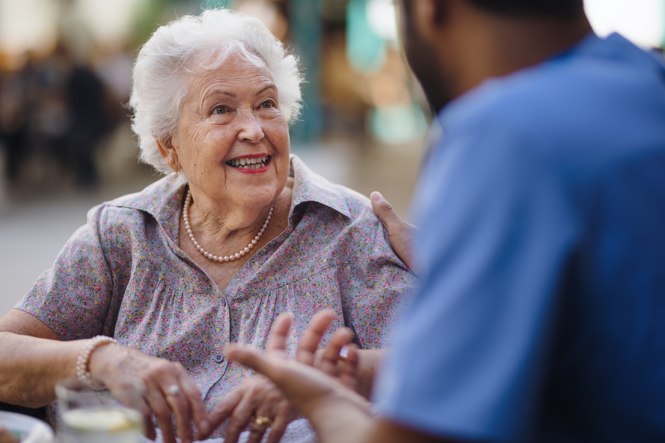 An elderly woman smiling warmly while talking with a caregiver outdoors, wearing a floral blouse and pearl necklace.