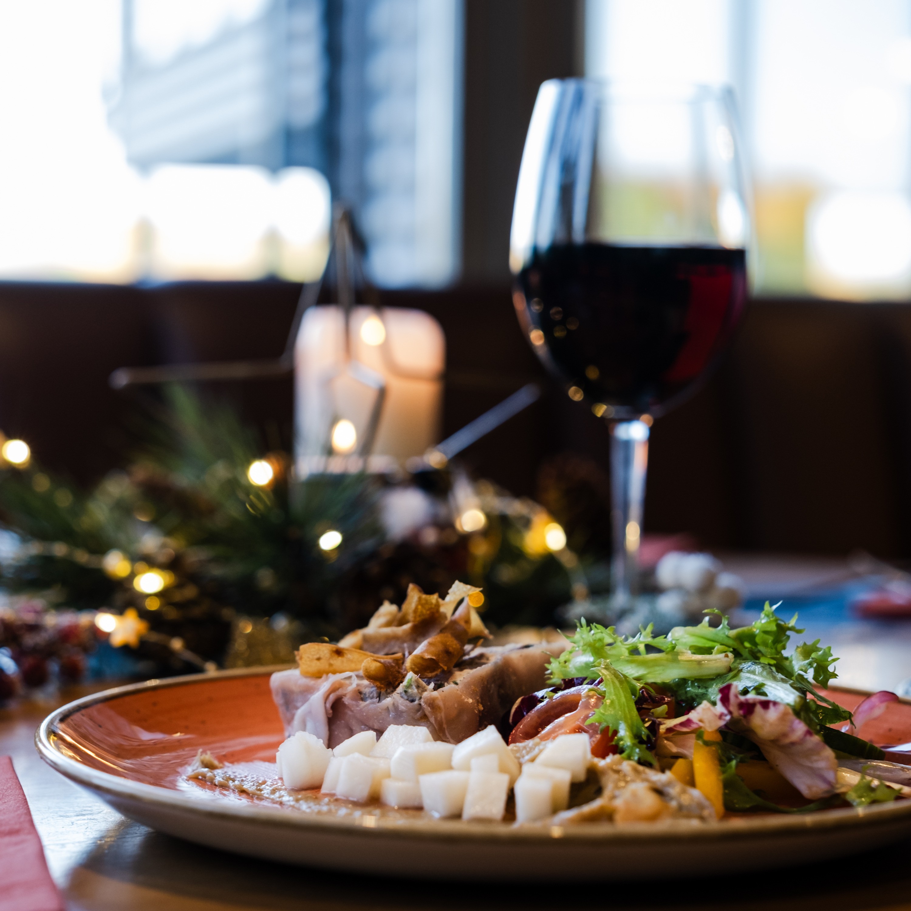 A festive dinner plate with roasted meat, salad, and diced vegetables, set beside a glass of red wine and holiday decorations with soft candlelight.