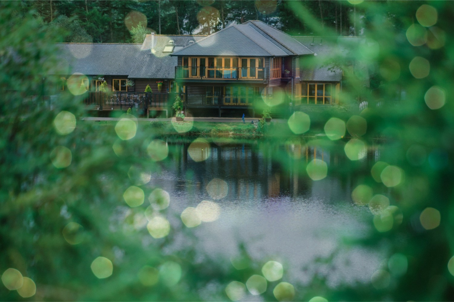 A cosy lakeside cabin with large windows is reflected in the calm water, framed by soft green bokeh lights and surrounding trees.