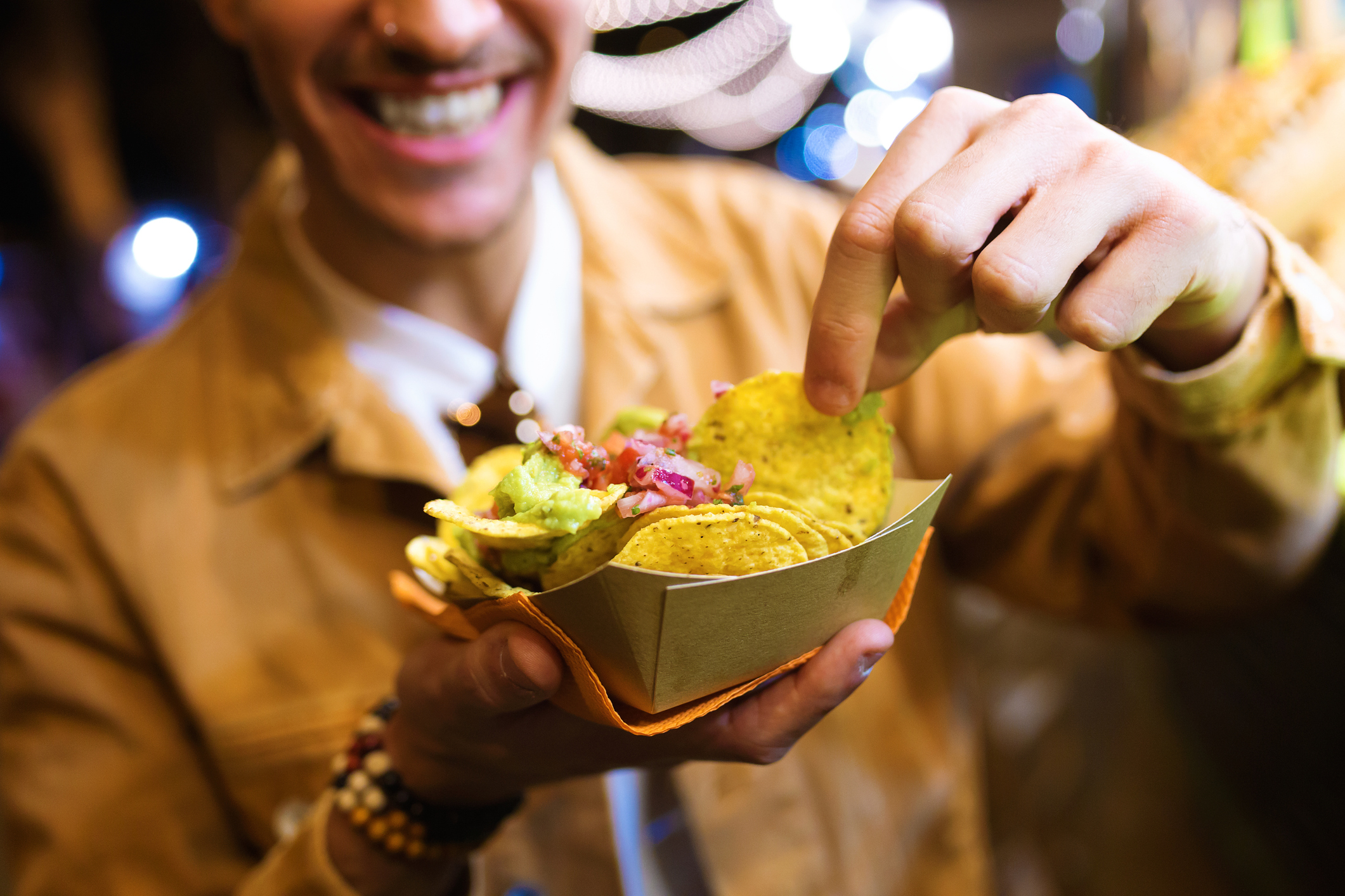 Smiling person holding a tray of nachos topped with guacamole and salsa, picking up a chip under warm lighting at what appears to be a festive event.