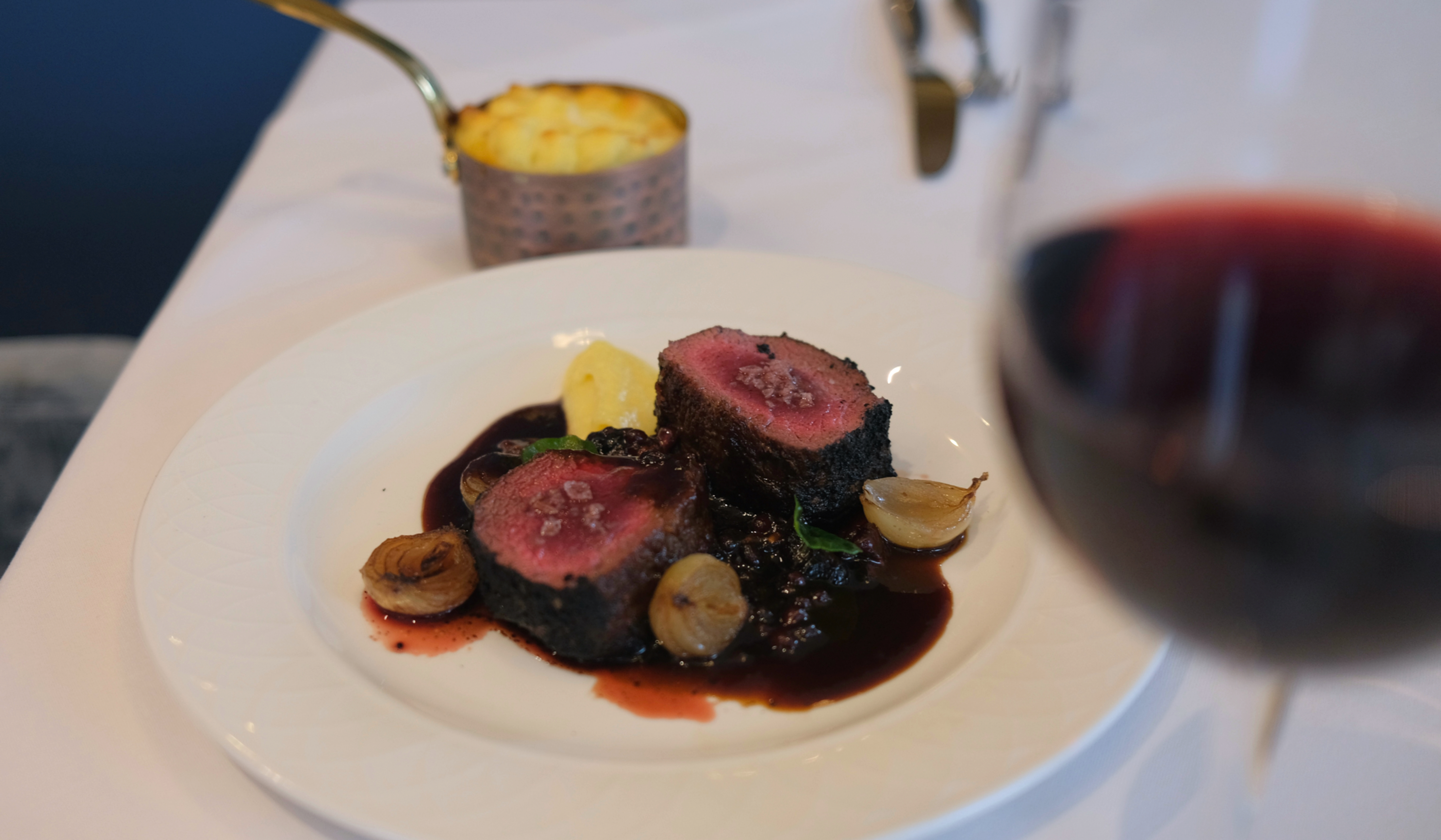 Plated venison dish at Gonville Kitchen with a red wine reduction, pearl onions, purée, and a copper pot of gratin, seen through a glass of red wine.