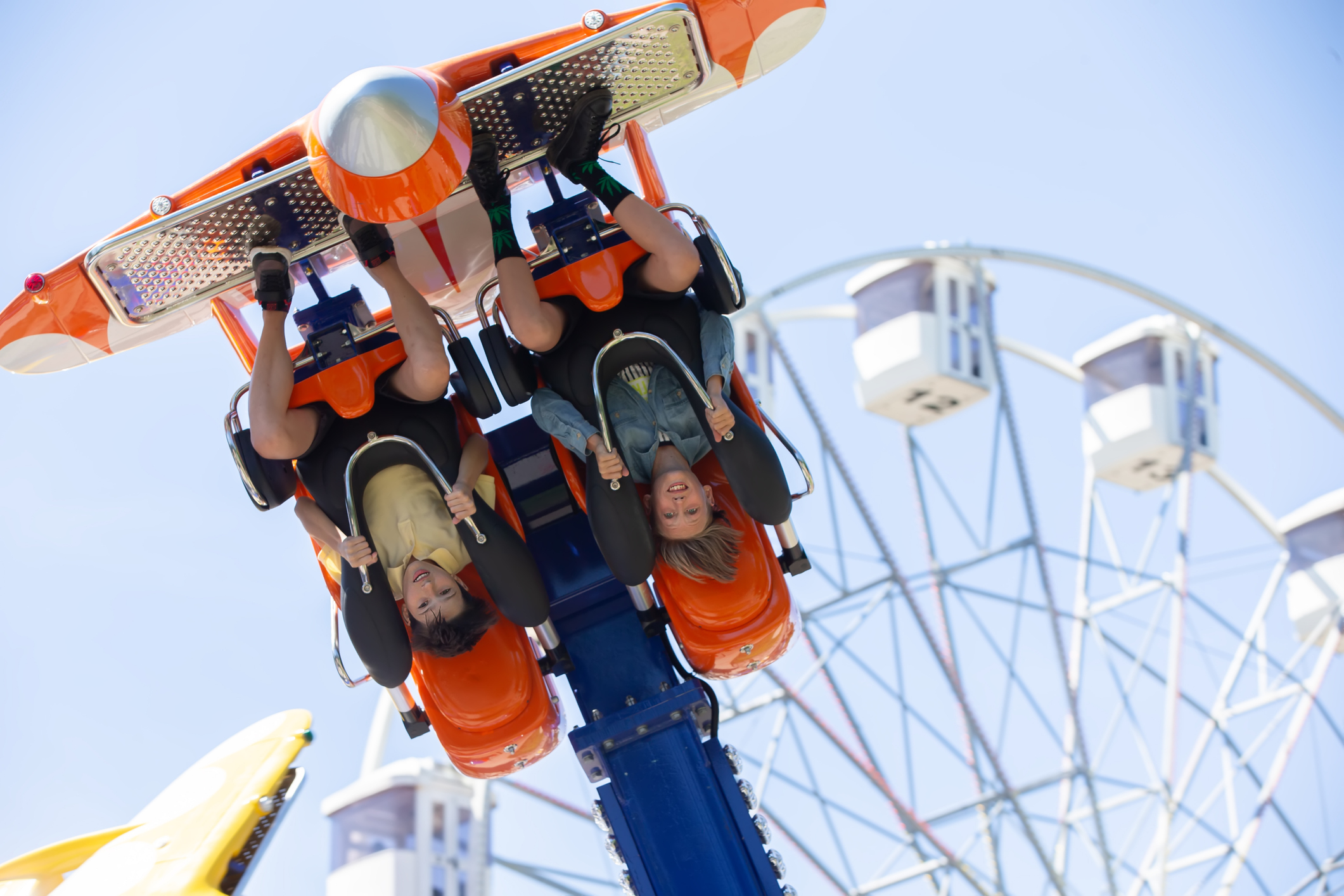 Two riders hang upside down on an orange amusement ride, smiling as a Ferris wheel blurs in the background against a clear blue sky.