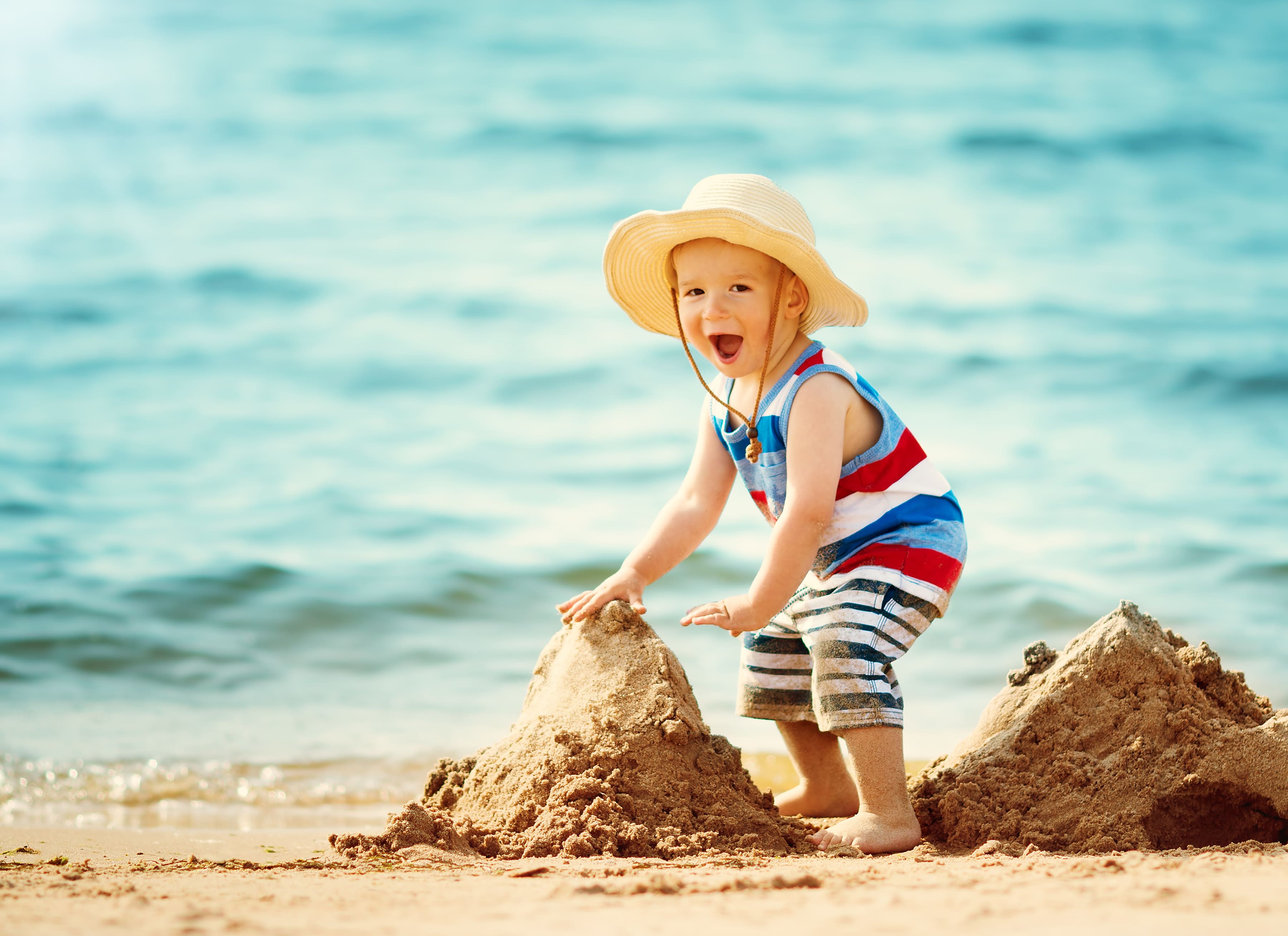 Smiling toddler in a sunhat builds a sandcastle on the beach, with gentle blue ocean waves in the background.