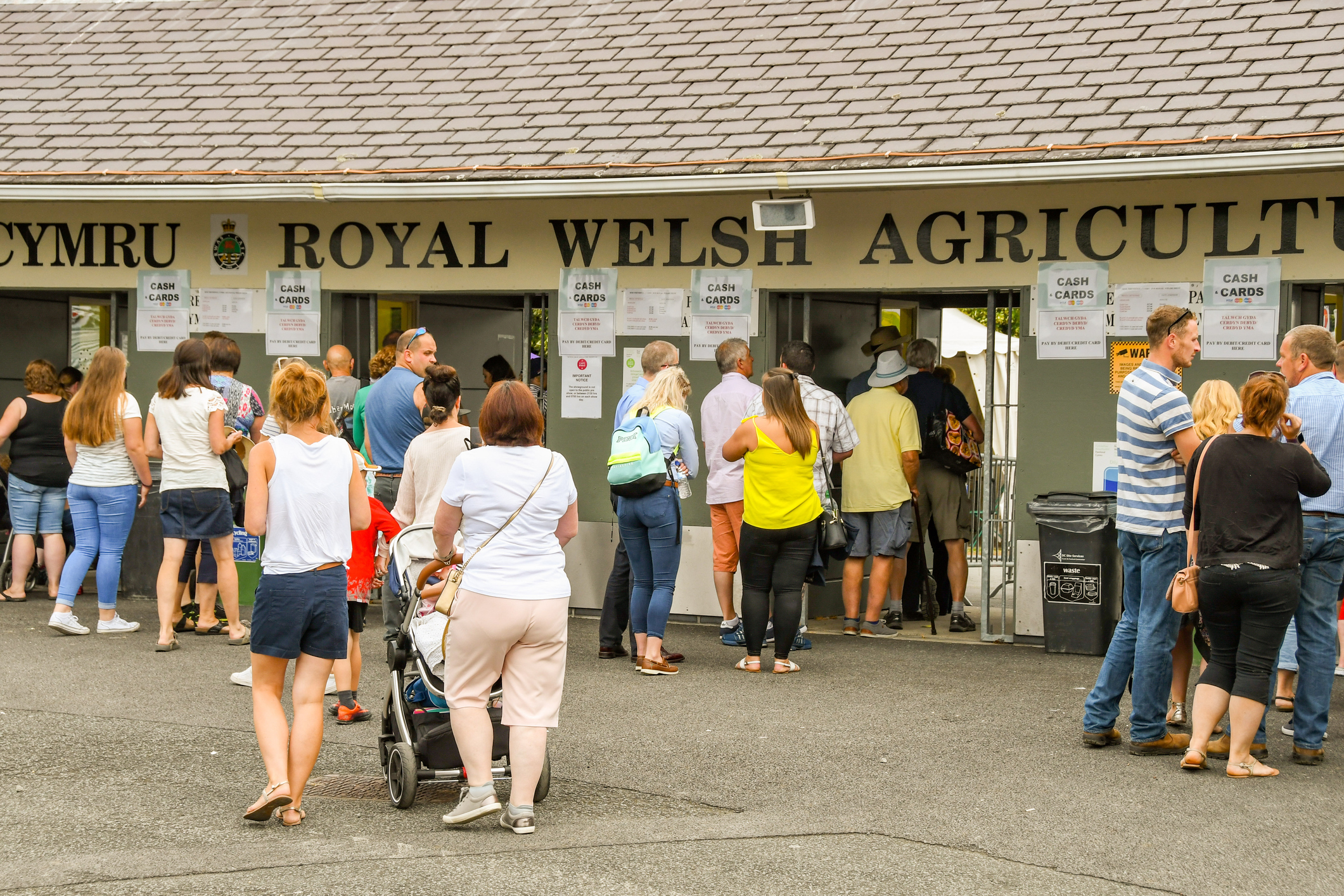 People queue at the entrance of the Royal Welsh Agricultural Show, with signs for ticket payment above and a mix of families and individuals waiting to enter.