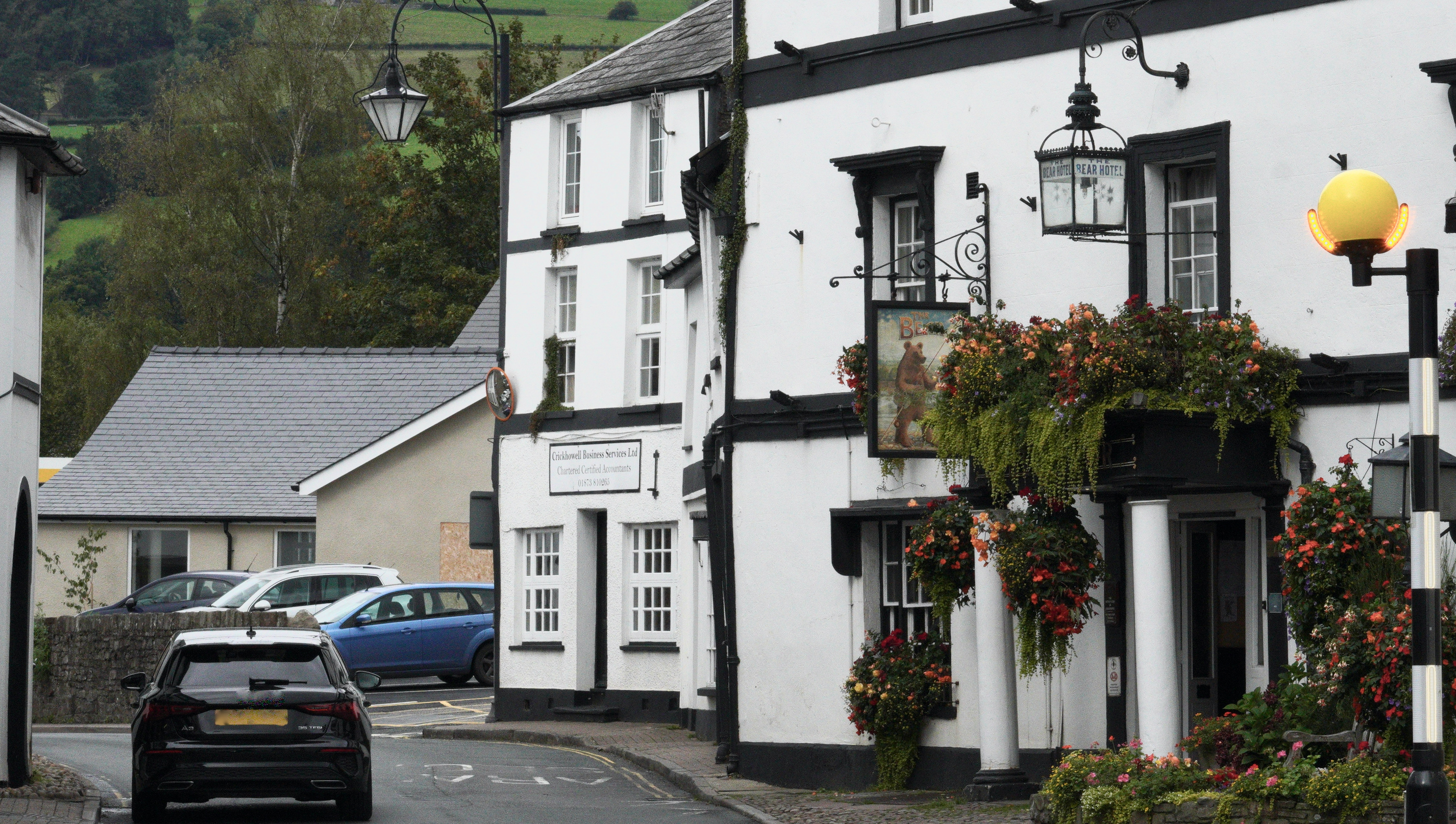 A charming view of Crickhowell High Street in Wales, featuring a black car driving past The Bear Hotel with flower-filled balconies and whitewashed buildings.
