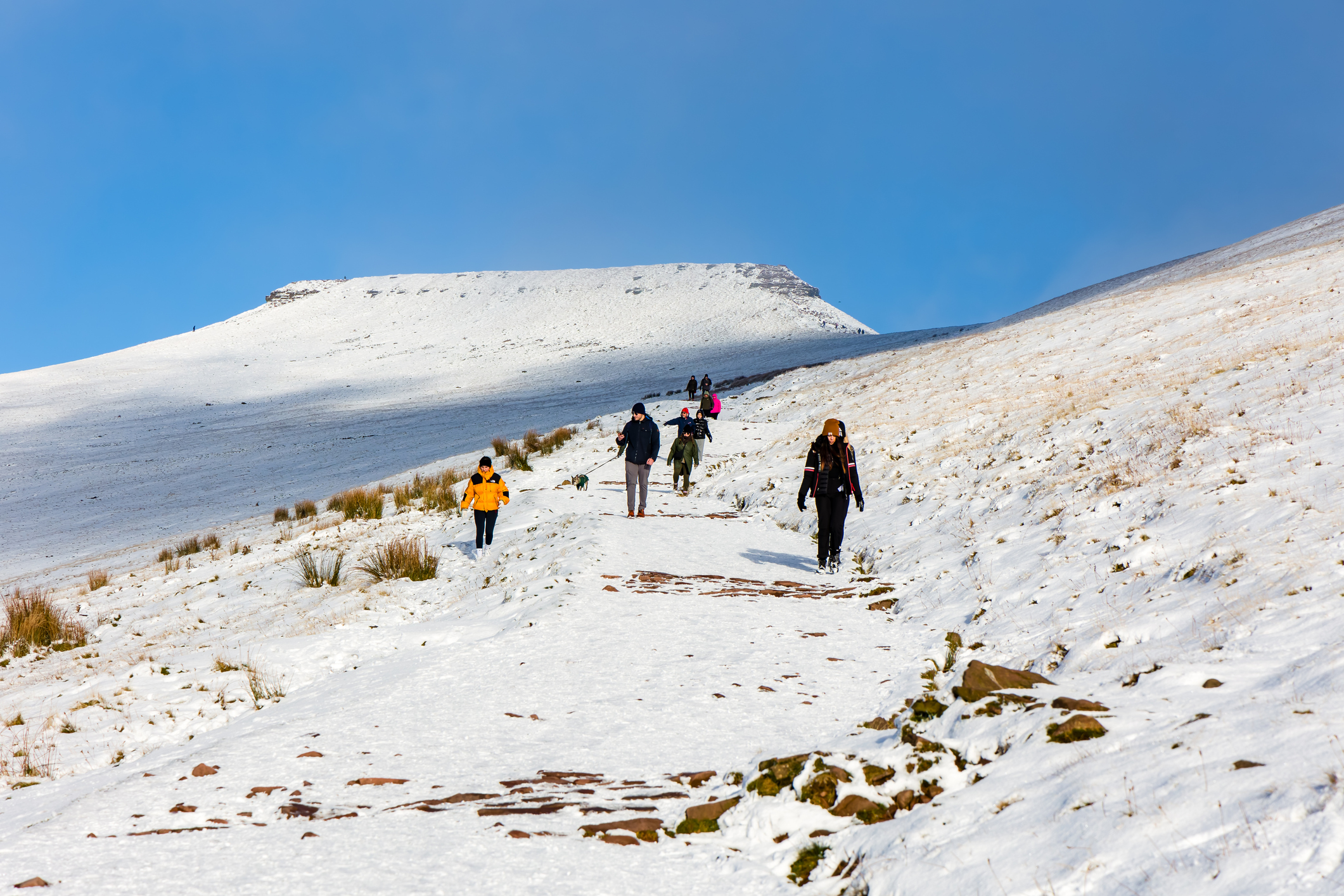 Hikers walk along a snowy trail on Pen y Fan in Bannau Brycheiniog, with clear skies and the snow-covered peak visible in the background.