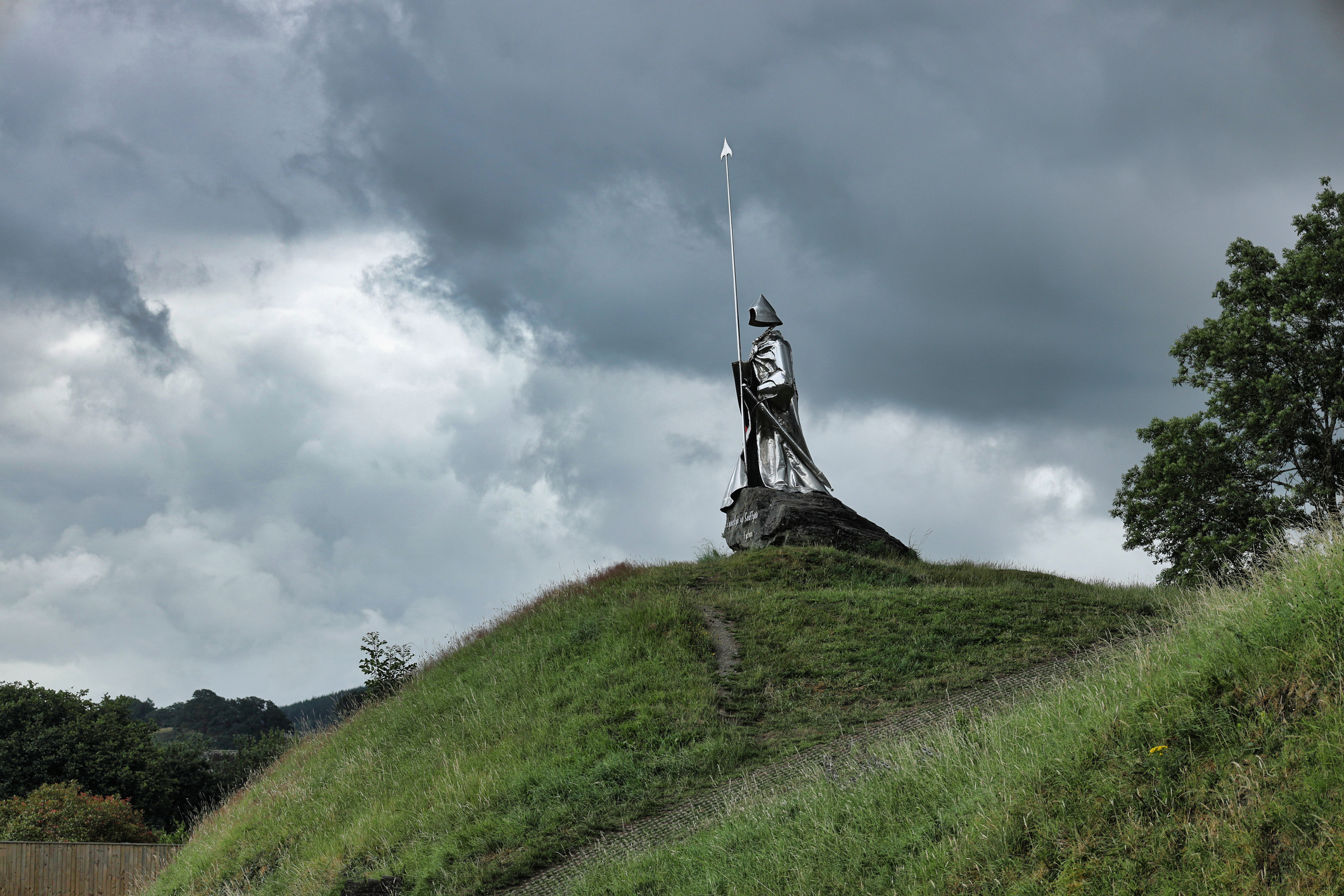 Monument to Llywelyn ap Gruffydd Fychan standing atop a grassy hill overlooking Llandovery, Carmarthenshire, beneath a dramatic cloudy sky.