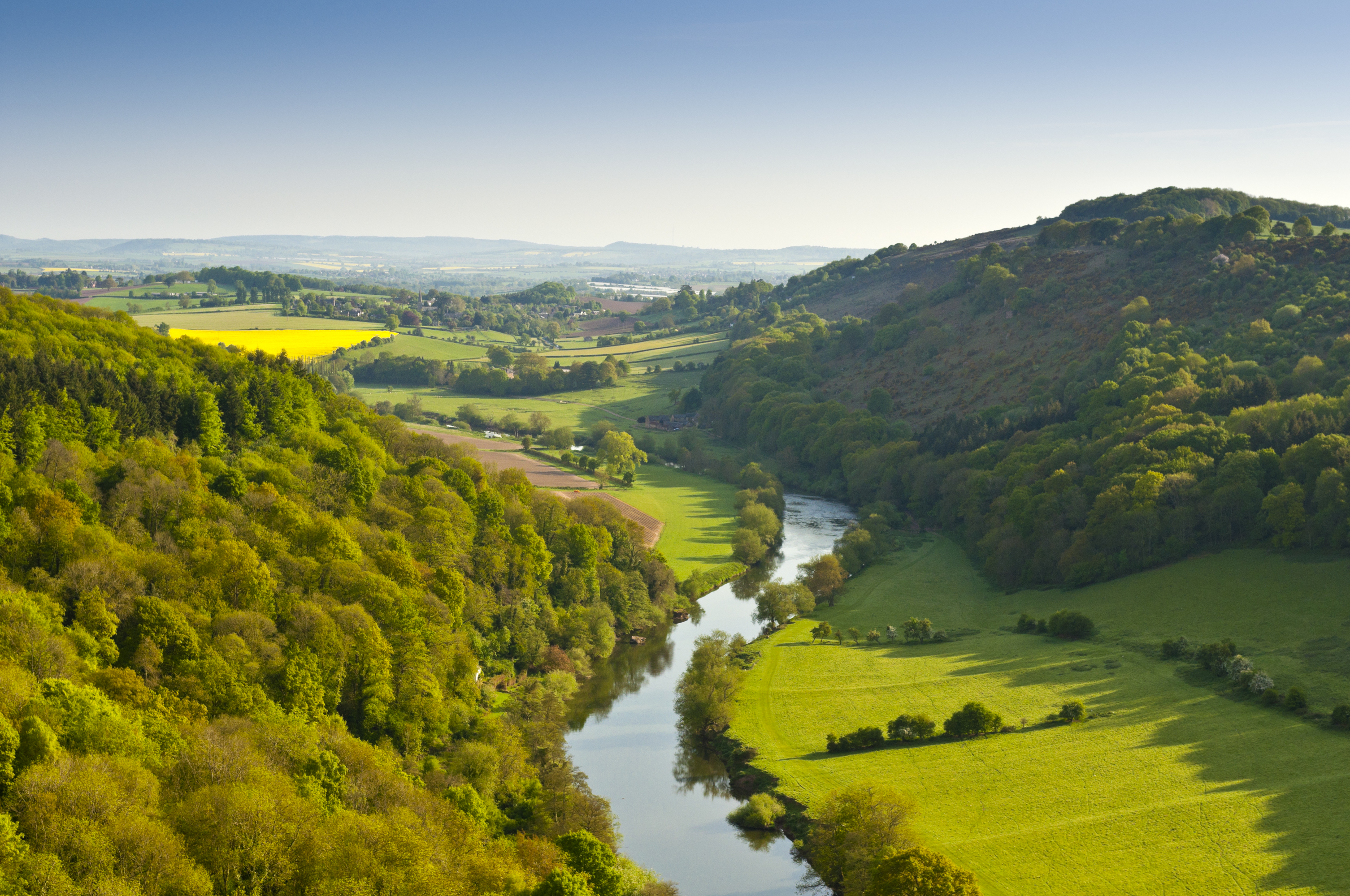 Aerial view of the Wye Valley in Wales, with a winding river cutting through lush green fields, rolling hills, and dense woodland under a clear blue sky.