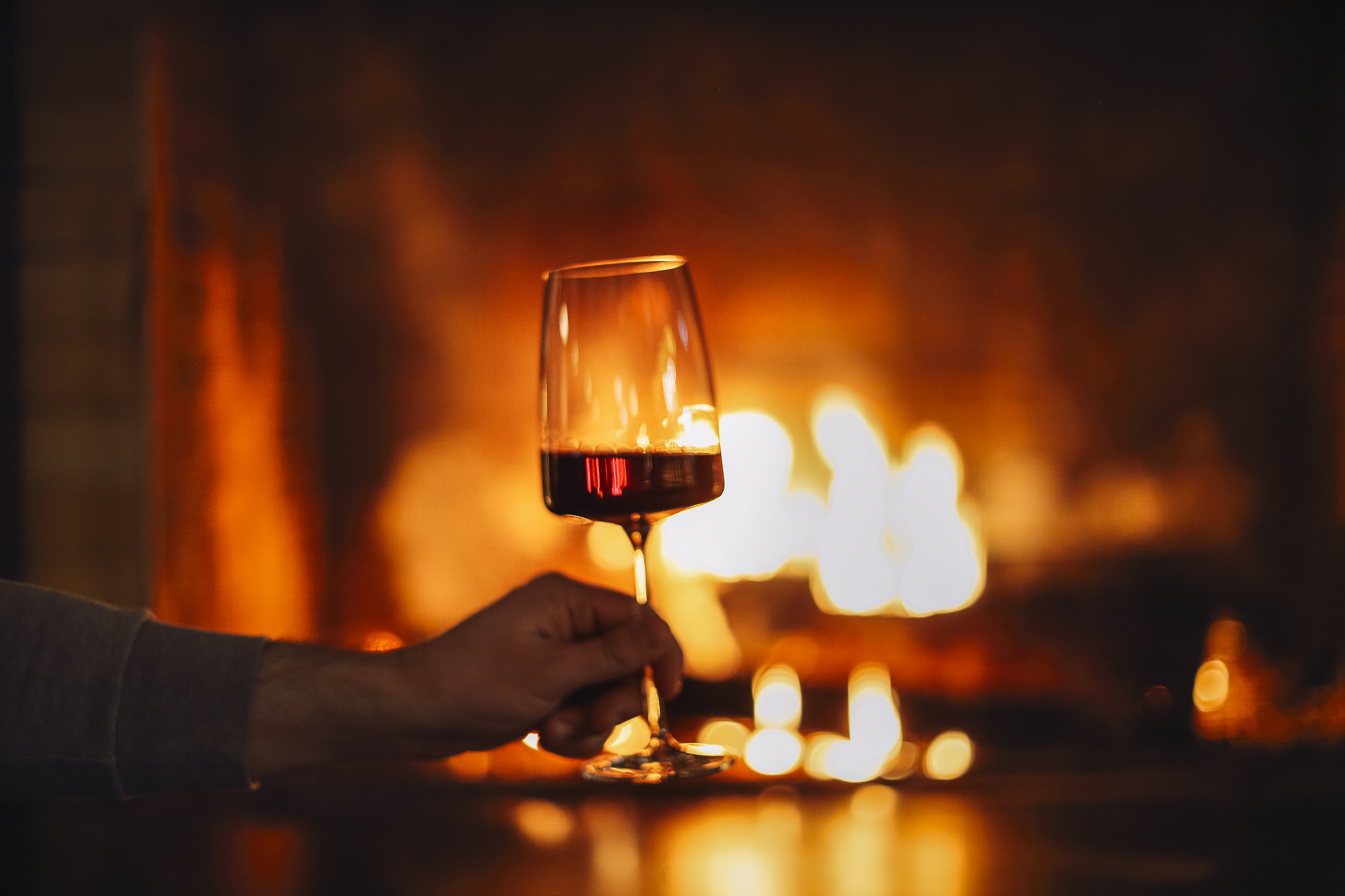 A hand holds a glass of red wine in front of a glowing fireplace, with warm flames casting a cosy, ambient light in the background.
