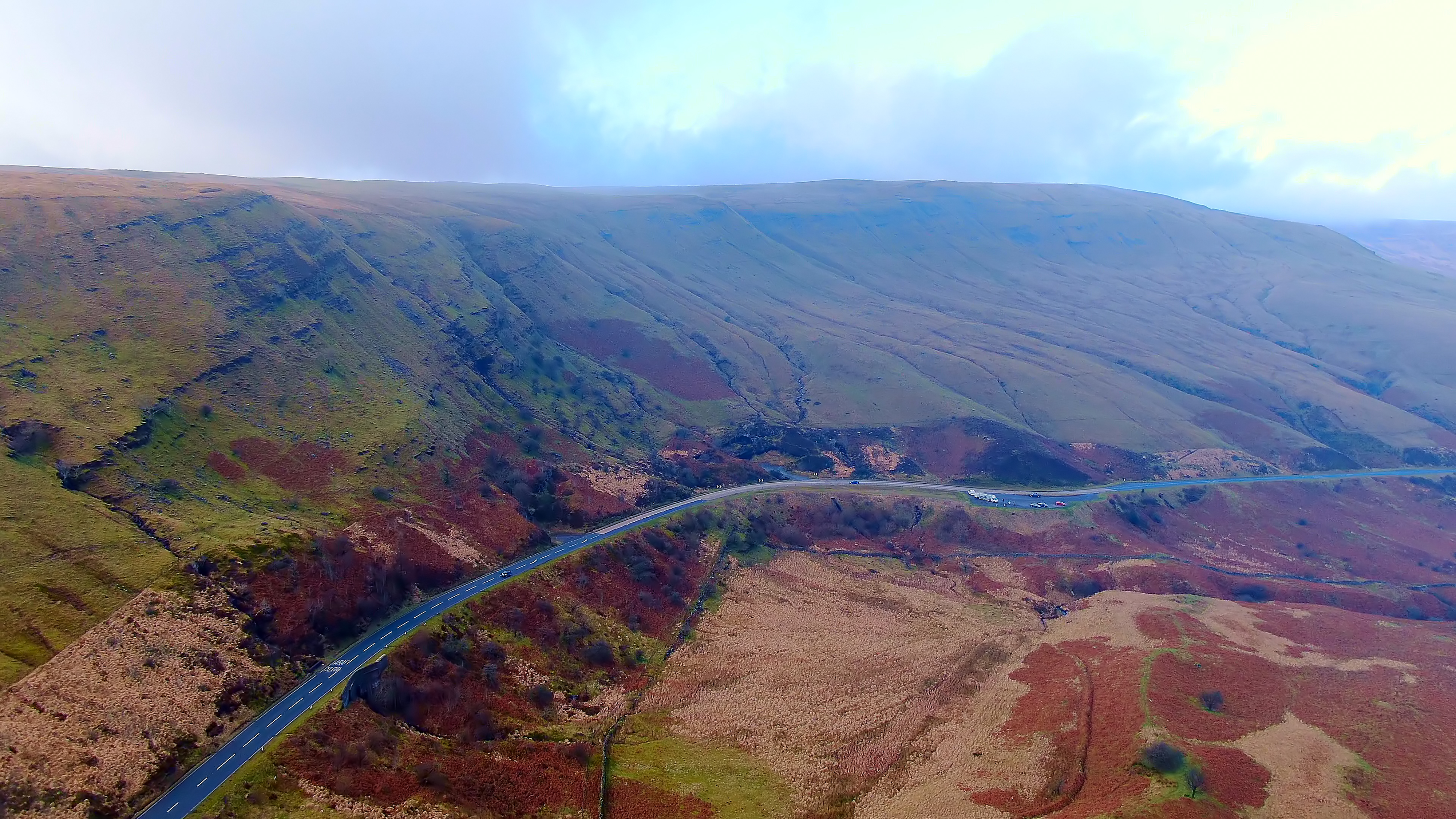 A winding road cuts through the rugged, mist-covered hills of Bannau Brycheiniog, with patchy grass, heather, and bracken stretching across the landscape.
