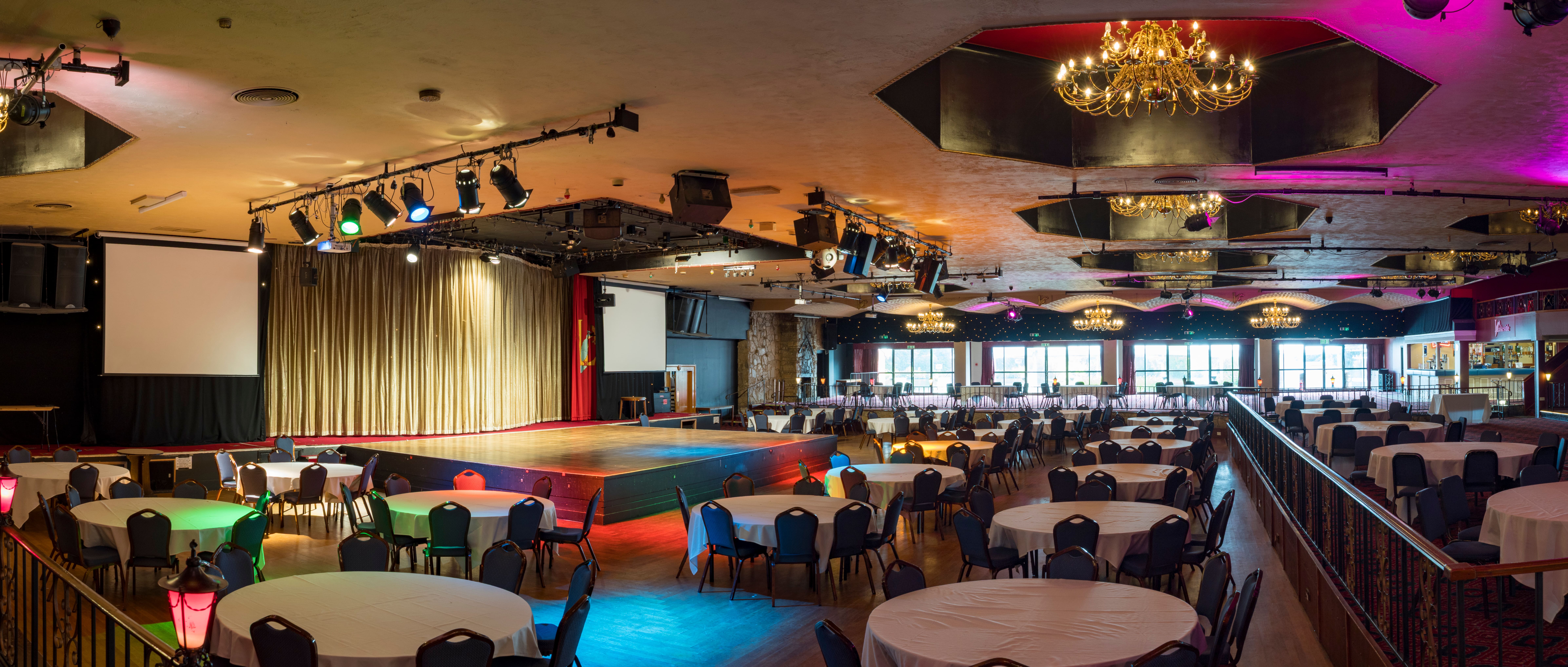 Main events hall at Lakeside Country Club in Frimley Green, Surrey, featuring a central stage, round banquet tables, chandeliers, and colourful lighting.