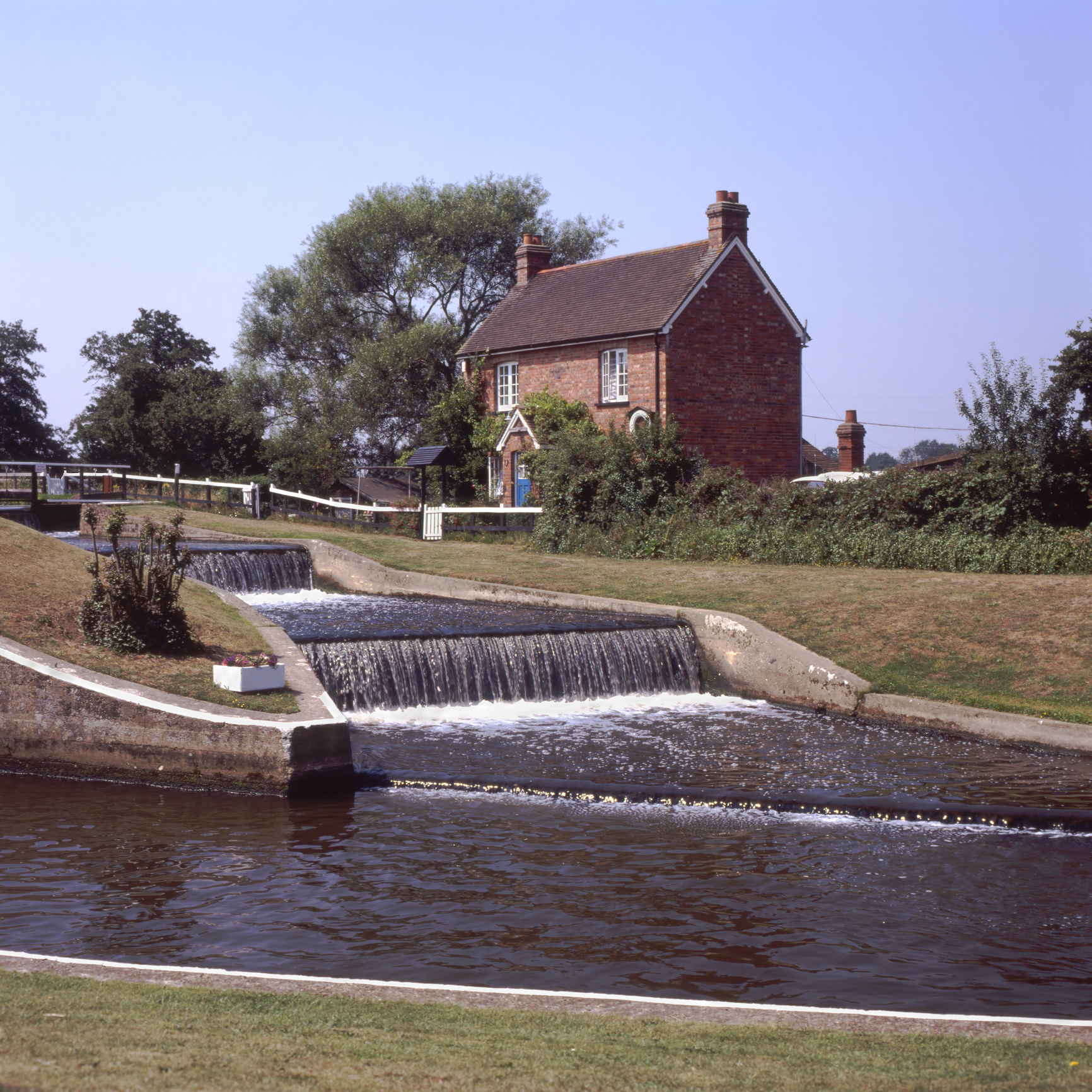 The weir at Ripley village in Surrey, with cascading water over concrete steps beside a red-brick cottage, greenery, and a clear blue sky in the background.