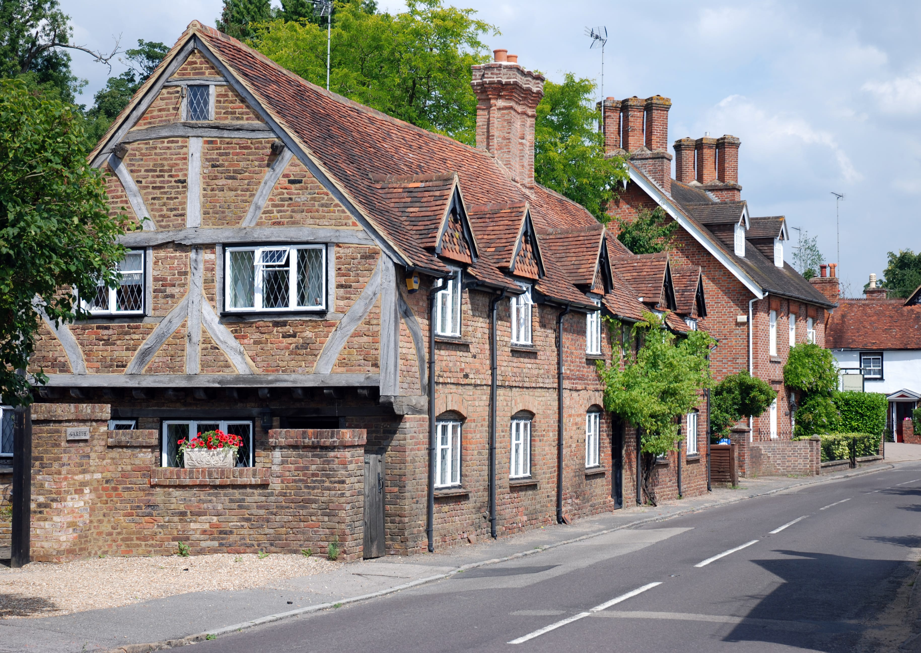 Historic brick and timber-framed cottages line Upper Street in Shere, Surrey, with red-tiled roofs, chimneys, and greenery along the quiet road.