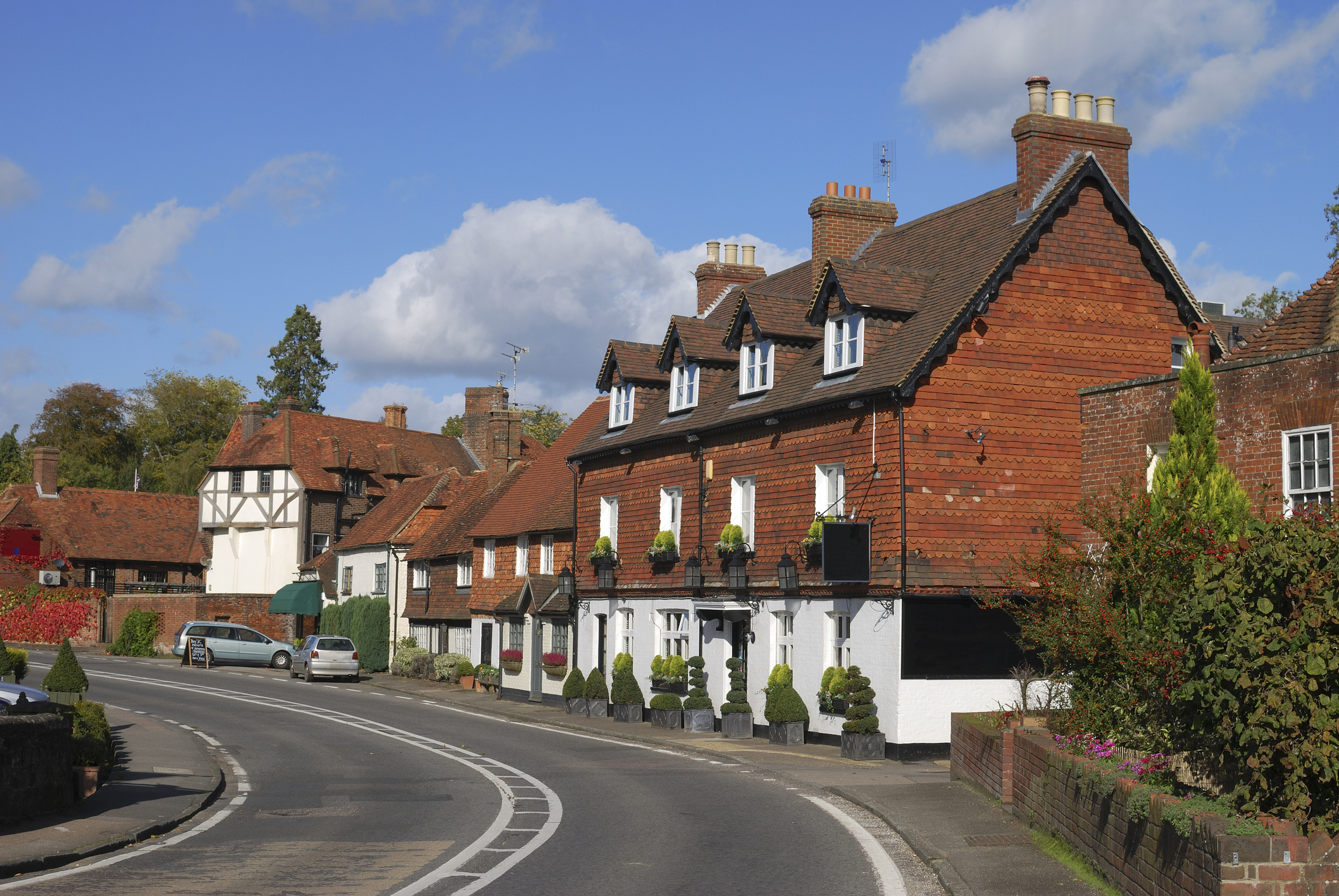 Curved street lined with historic red-tiled buildings in Chiddingfold, Surrey, under a bright blue sky with scattered clouds and well-kept greenery.