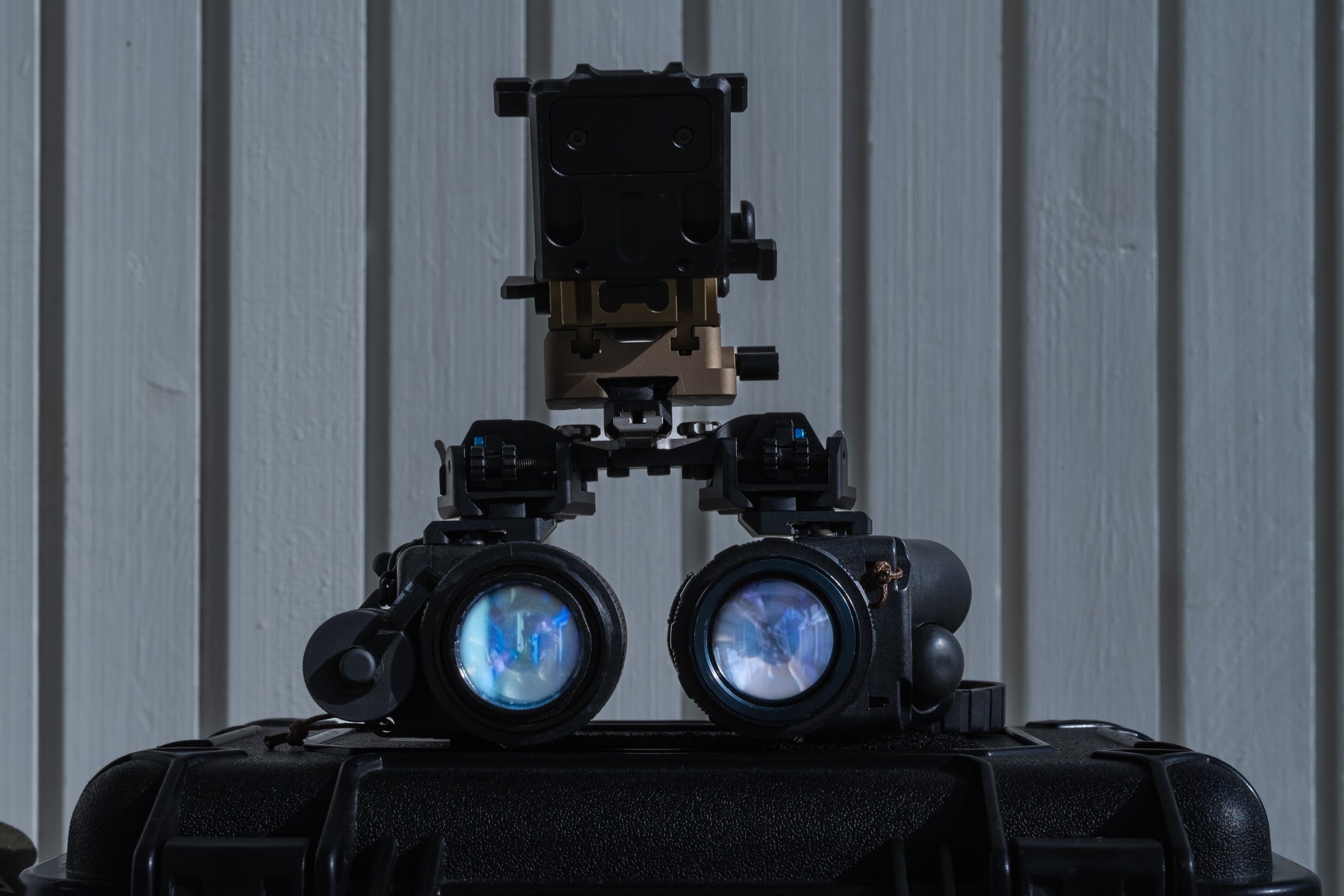 Night vision goggles with a helmet mount attachment resting on a hard protective case, positioned against a vertical wooden panel background.