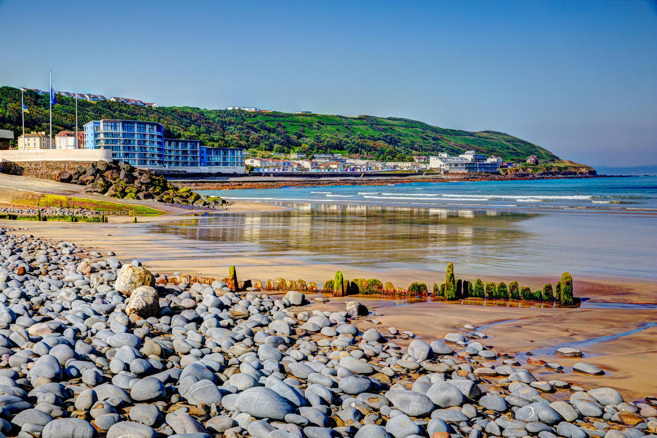 Pebble-covered beach at Westward Ho! near Bideford in North Devon, with calm waves, modern seafront buildings, and green hills under a clear blue sky.