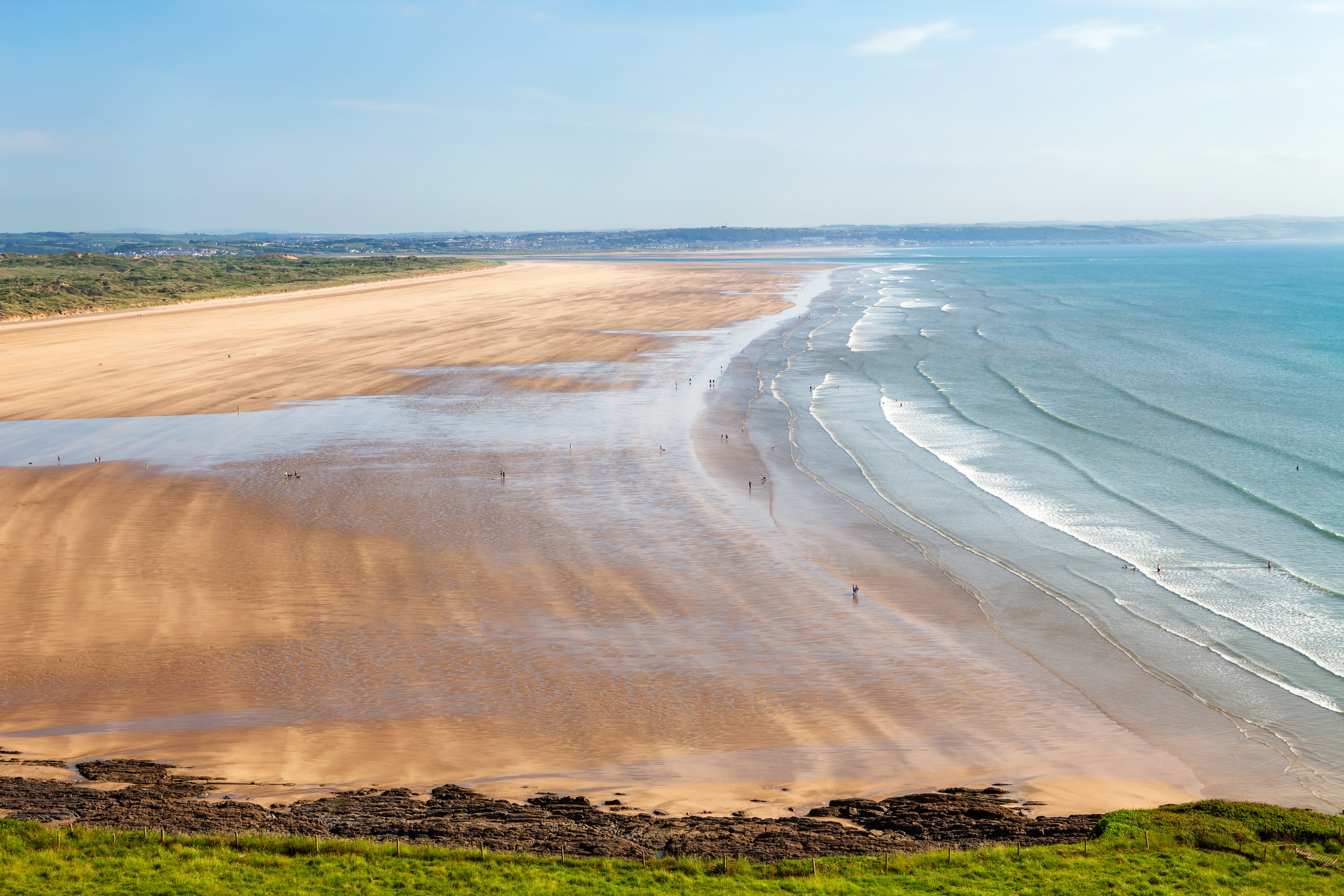 Wide view of Saunton Sands in North Devon, with gentle waves lapping a vast sandy beach, scattered visitors, and grassy cliffs in the foreground.