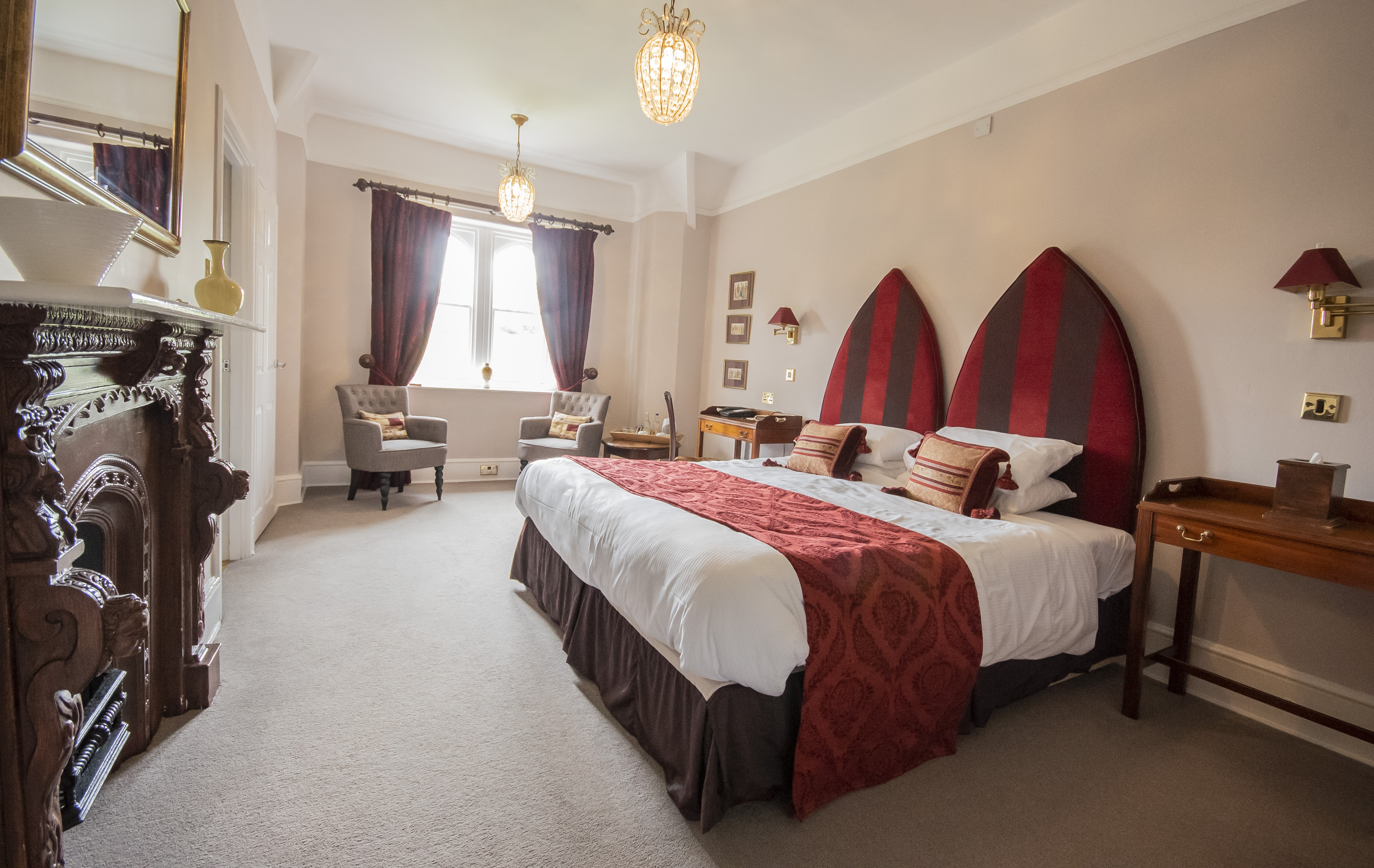 The Monkleigh Room at Northcote Manor, featuring a double bed with red and gold accents, elegant furnishings, armchairs by a window, and a carved fireplace.