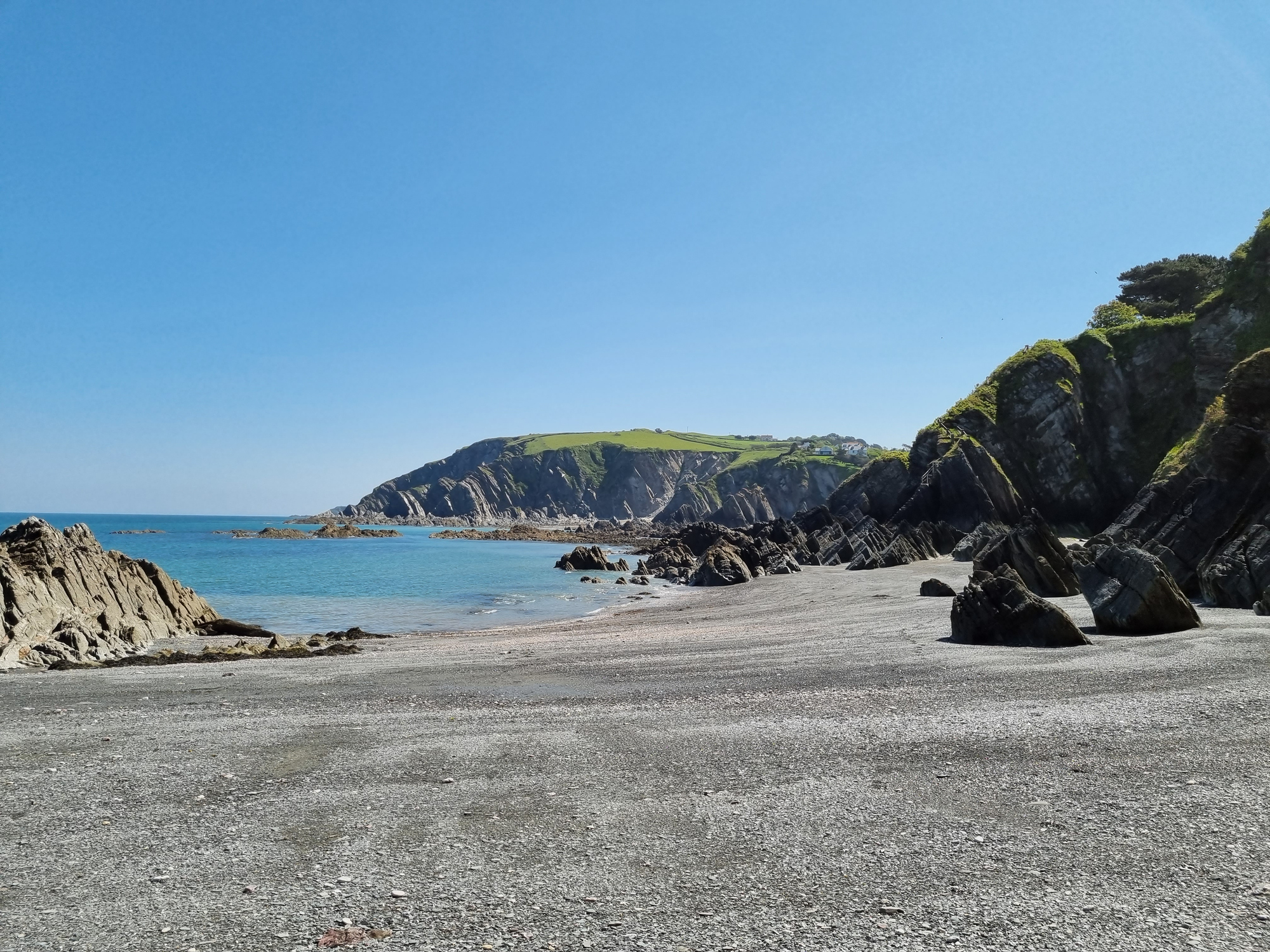 Rocky shoreline at Lee Bay in North Devon, with jagged cliffs, calm turquoise water, and a clear blue sky on a bright sunny day.