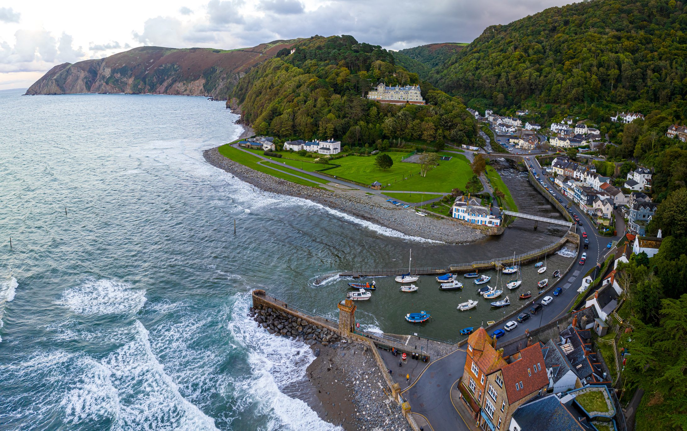 Aerial view of Lynmouth, showing the harbour, boats, and pebble beach where the river meets the sea, surrounded by green hills and coastal cliffs.