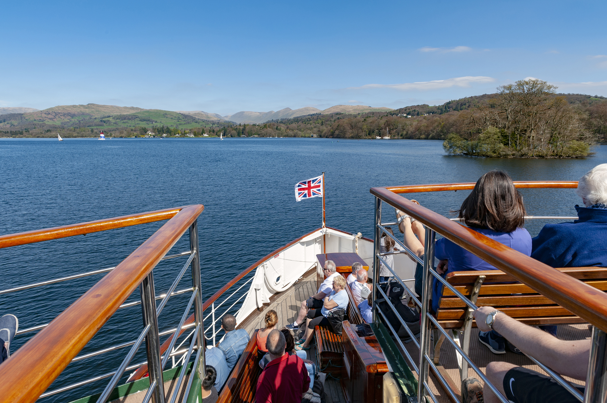 Passengers relax on a boat deck cruising a lake under a clear blue sky, with a Union Jack flag at the bow and scenic hills in the background.
