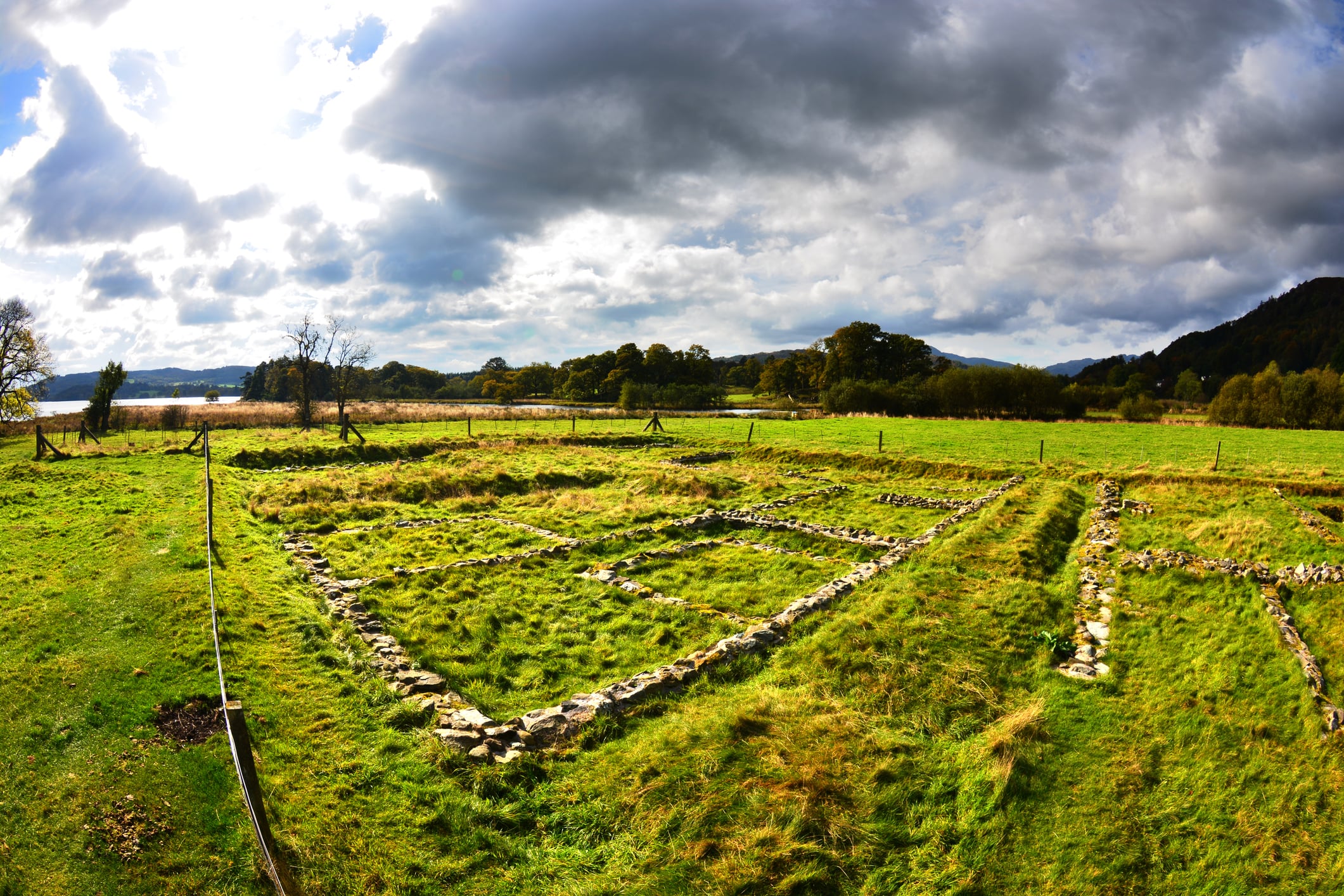 Stone foundations mark the remains of a Roman settlement at Waterhead, set in a green field under dramatic clouds with trees and hills in the distance.