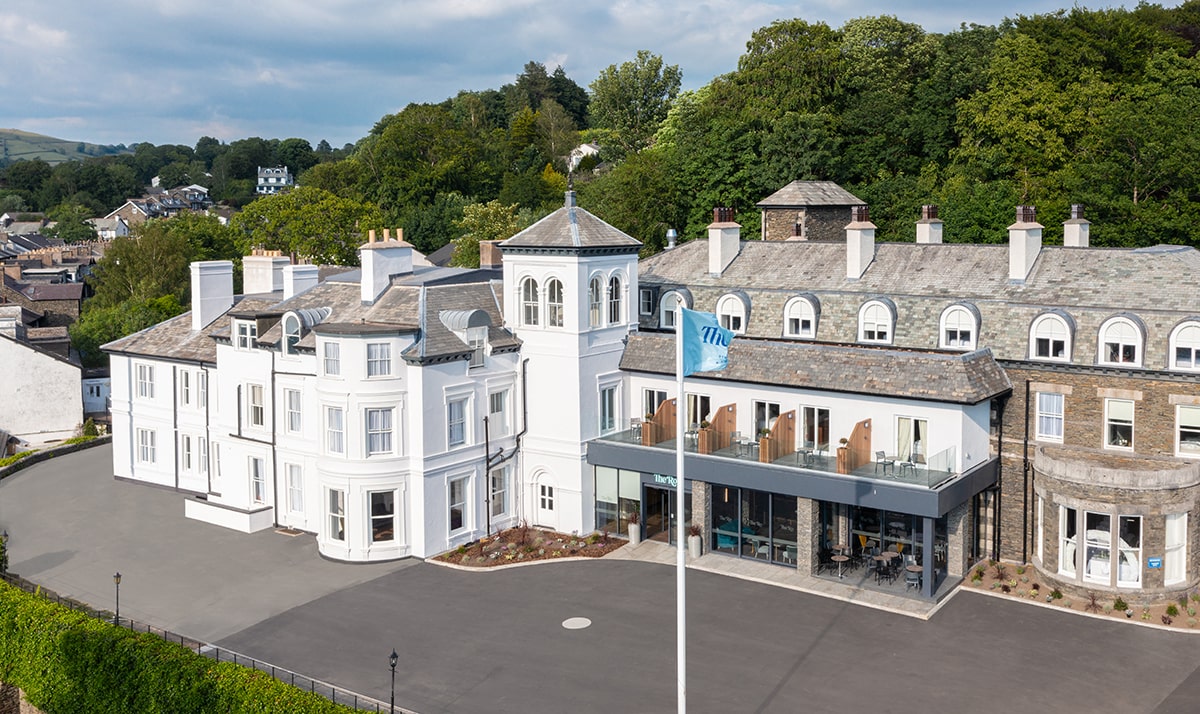 The Ro Hotel in Bowness-on-Windermere, a large white and stone-fronted building with a tower, balconies, and modern glass-fronted café, set near woodland.