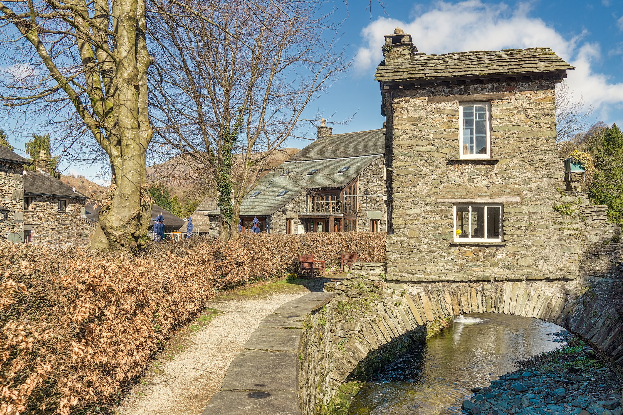 The Bridge House in Ambleside, a small stone building straddling a stream, with a footpath, bare trees, and traditional slate-roofed cottages nearby.
