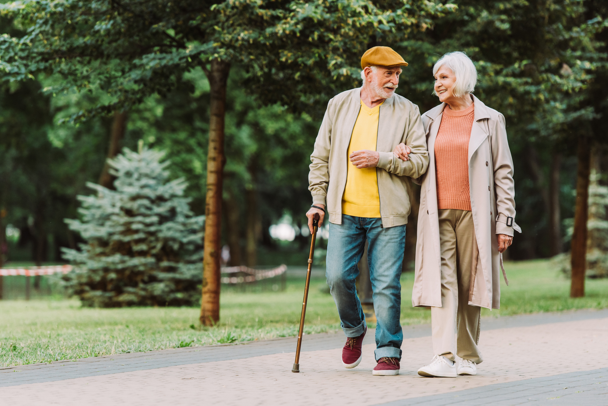 An elderly couple walks arm in arm through a park, smiling at each other. The man uses a cane and both wear light jackets and casual clothes.