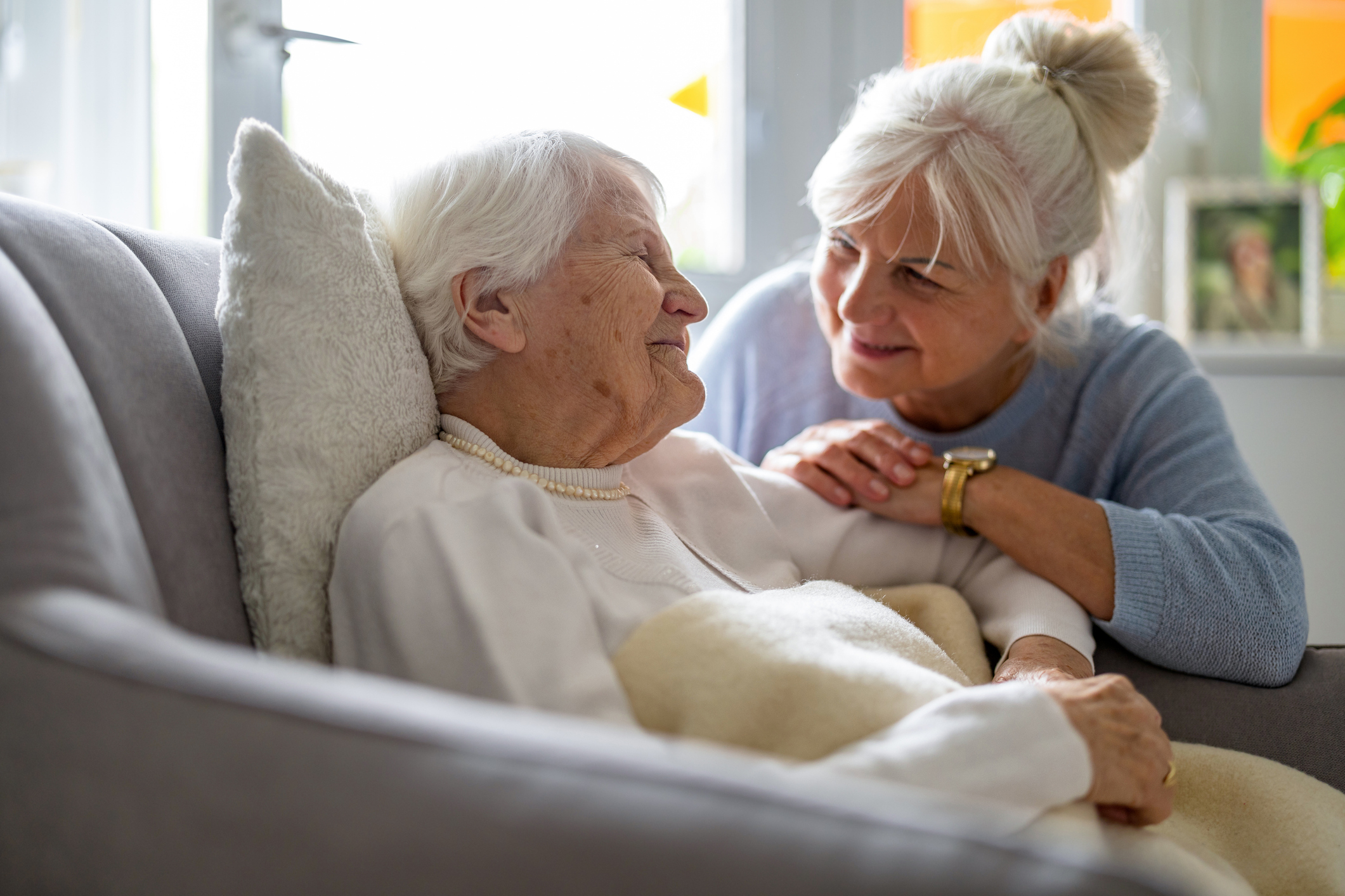 An elderly woman lies on a couch smiling warmly at another older woman who is sitting beside her, holding her arm and leaning in affectionately.