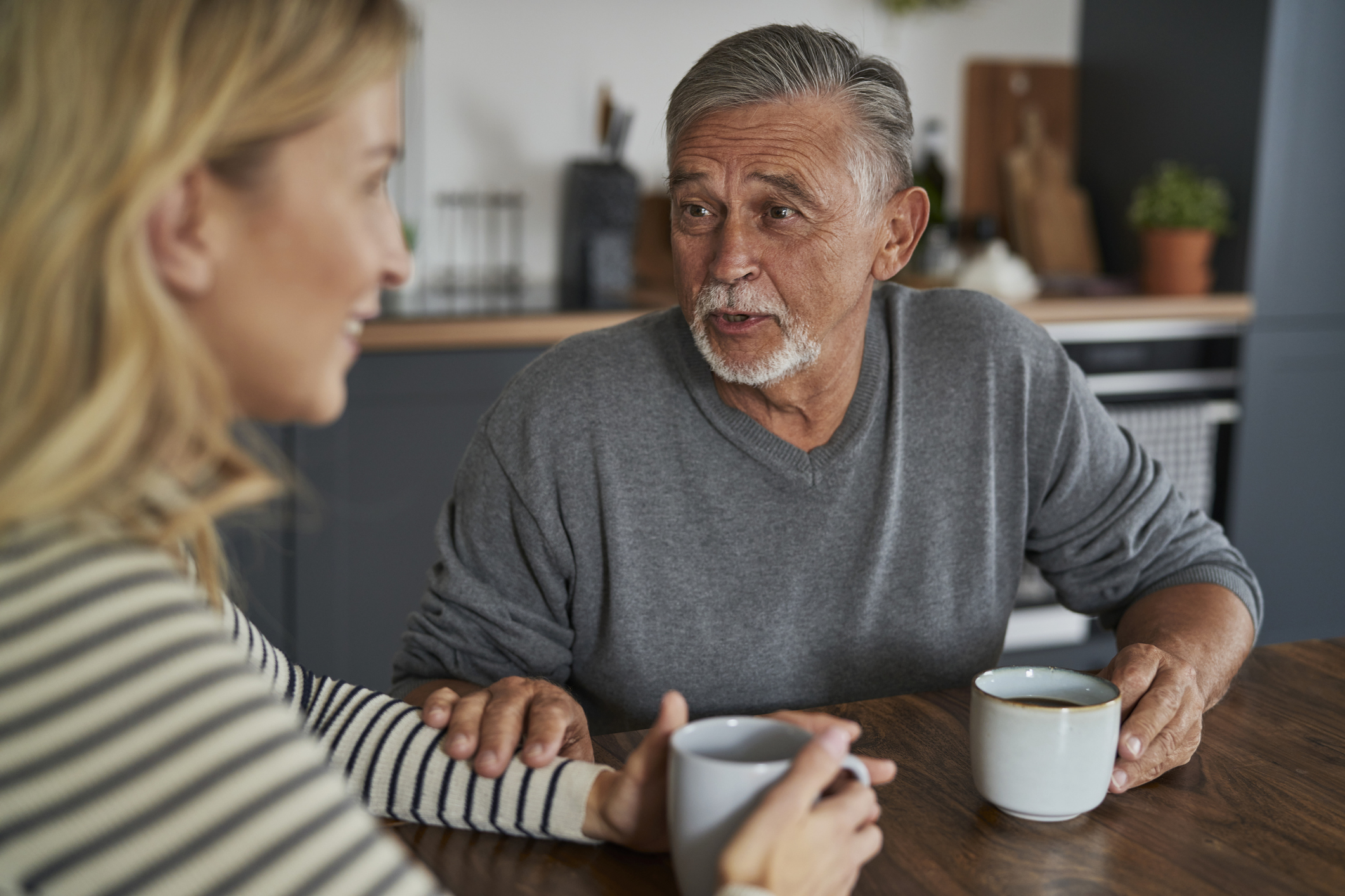 An older man and a younger woman sit at a table with mugs, having a warm conversation. The man gently touches her arm while speaking.