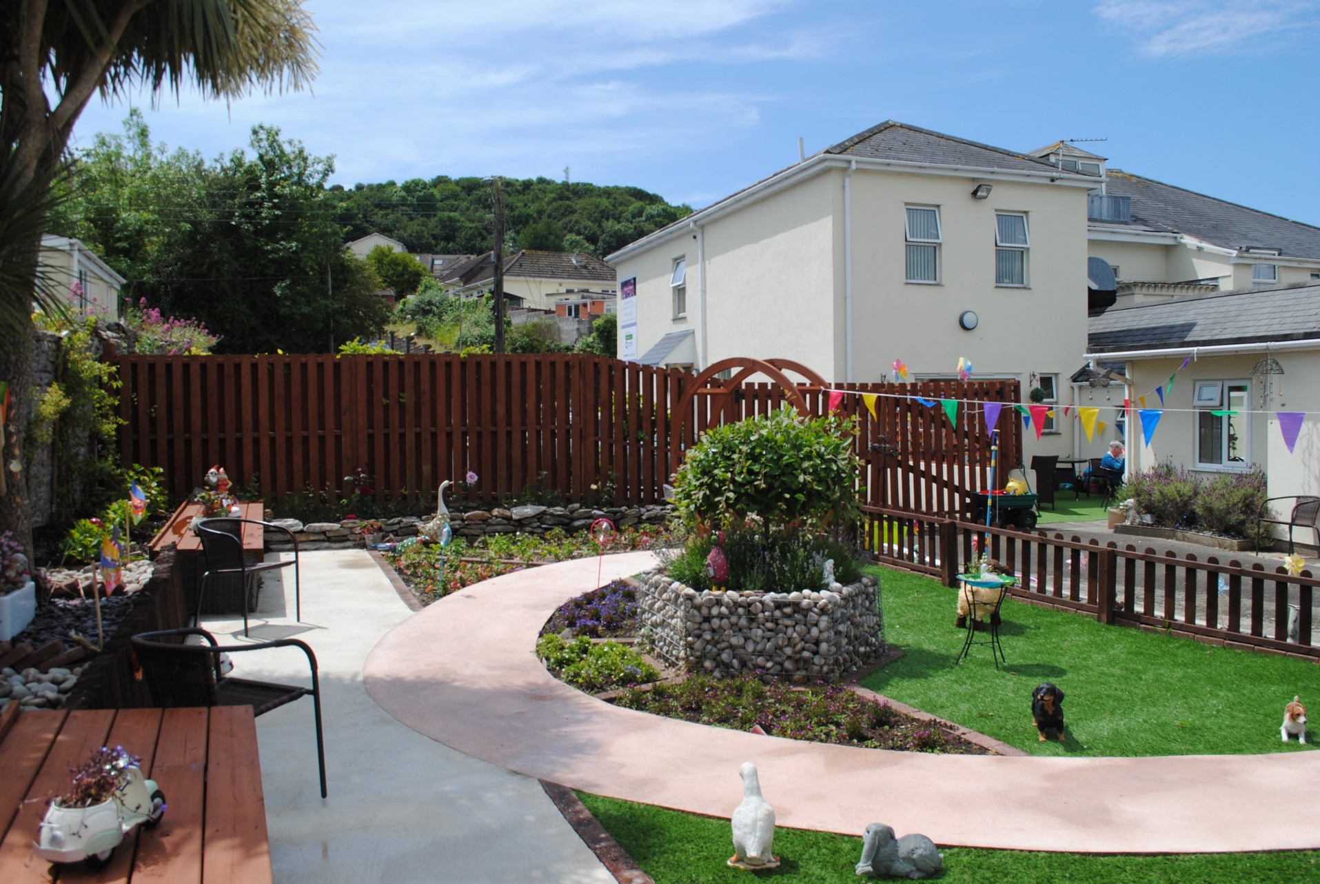 Sunny garden at Wisteria House Plymstock with a winding path, flower beds, benches, bunting, and decorative animal ornaments outside a cream-colored house.