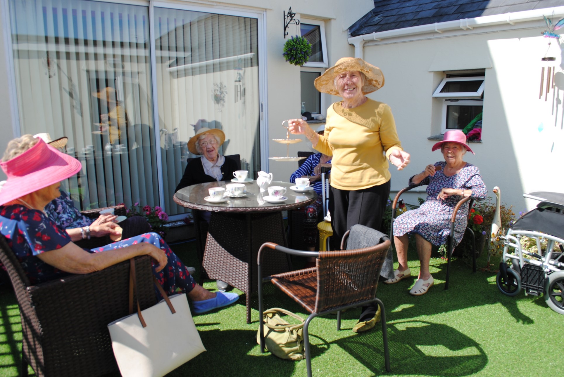 Elderly women in colourful hats enjoying a sunny tea party in the garden of Wisteria House’s Plymstock home, gathered around a table with teacups.